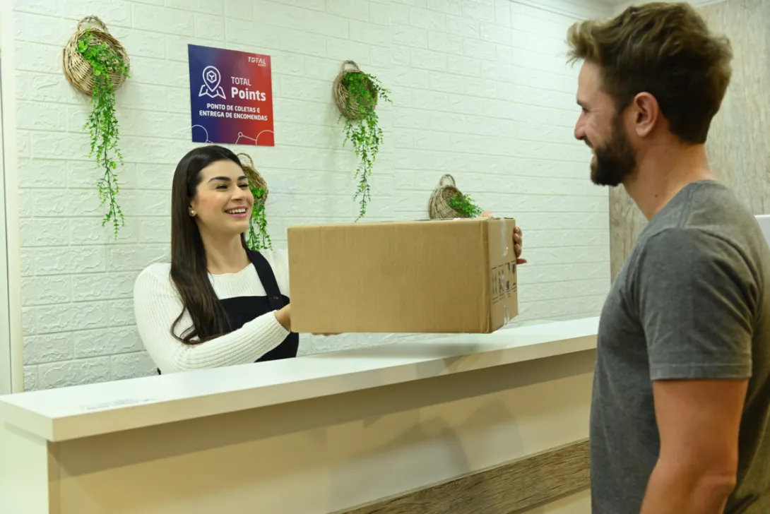 Uma recepcionista entrega um pacote de papelão a um homem sorridente do outro lado do balcão. A área da recepção tem uma parede de tijolos brancos com duas decorações de plantas penduradas e uma placa “Total de Pontos”. Ambos os indivíduos parecem estar em uma interação alegre.