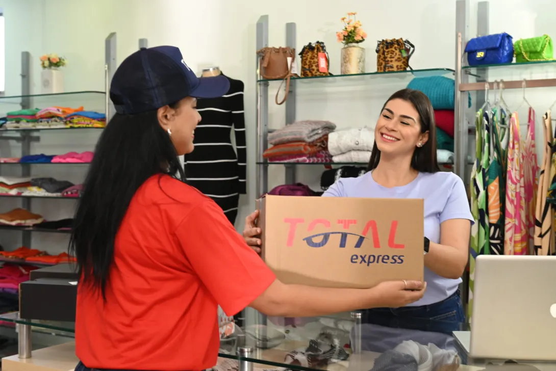 Um entregador de camisa vermelha e boné azul-marinho entrega um pacote a uma mulher atrás do balcão de uma loja de roupas. O pacote diz “Total Express”. A loja possui prateleiras com roupas dobradas, bolsas e itens diversos ao fundo.