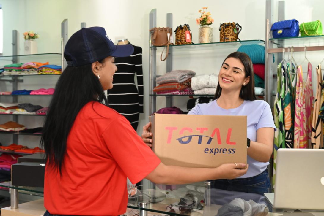 Um entregador de camisa vermelha e boné azul-marinho entrega um pacote a uma mulher atrás do balcão de uma loja de roupas. O pacote diz “Total Express”. A loja possui prateleiras com roupas dobradas, bolsas e itens diversos ao fundo.