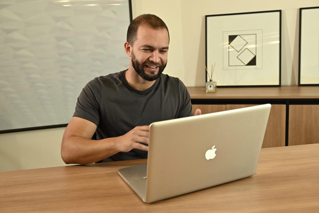 Um homem com barba sorri enquanto usa um laptop em uma mesa. Há estampas geométricas emolduradas e um difusor de palheta no fundo.