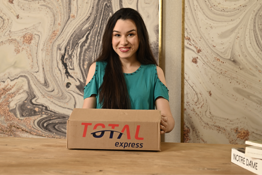 Uma mulher com longos cabelos escuros está sorrindo e segurando uma caixa de papelão com a inscrição "TOTAL express" enquanto está sentada em uma mesa de madeira. O fundo apresenta um desenho de parede em mármore. Um livro com o título "NOTRE DAME" está parcialmente visível na mesa à direita.