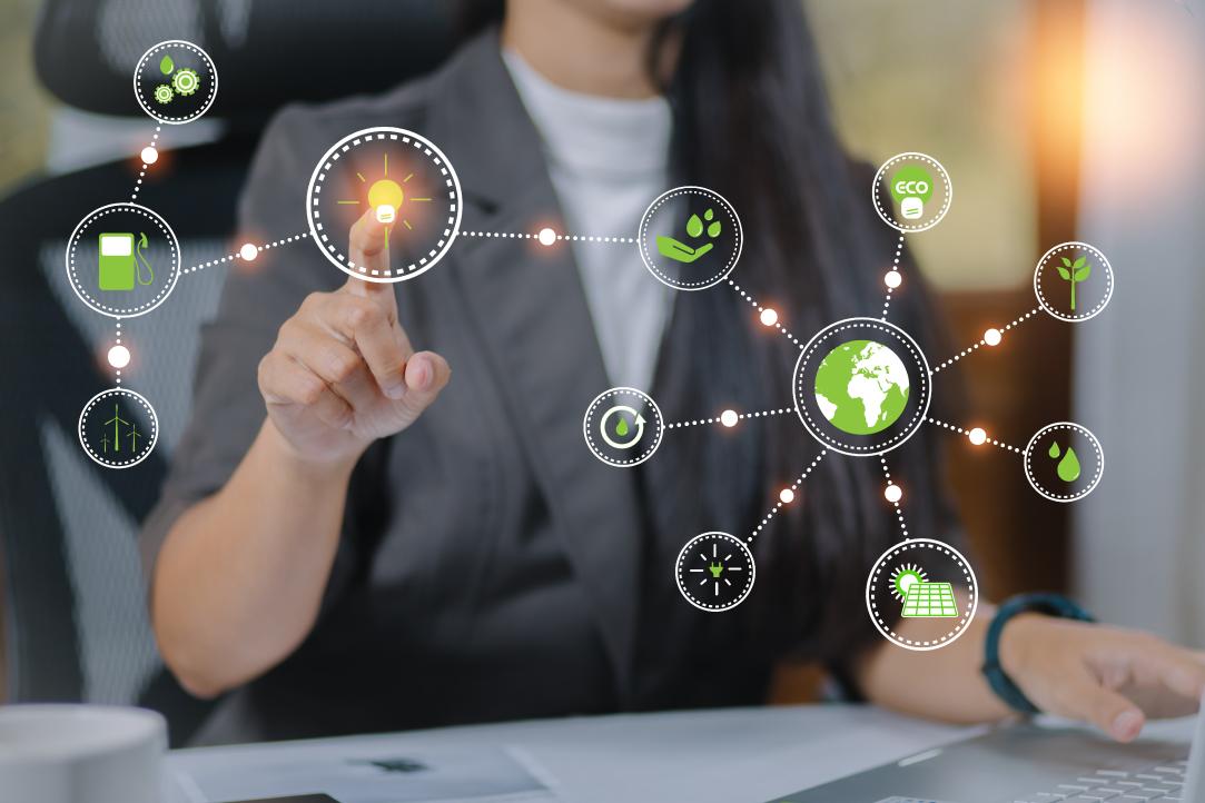 Uma empresária de terno formal toca um ícone iluminado em uma interface virtual que representa a sustentabilidade ambiental. Vários ícones verdes conectados a cercam, incluindo símbolos ecológicos e um globo. Ela está trabalhando em uma mesa com um computador.