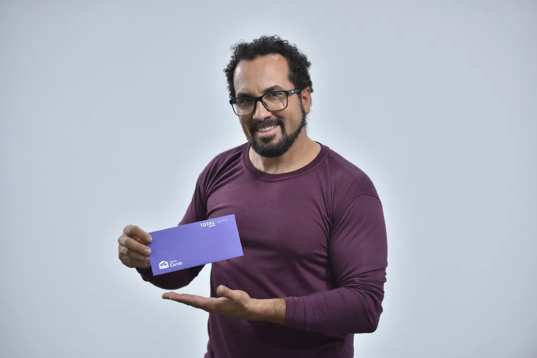 Um homem de cabelos cacheados, barba e óculos, vestindo uma camisa marrom de mangas compridas, segura e aponta para um envelope roxo com um sorriso. O fundo é totalmente branco.