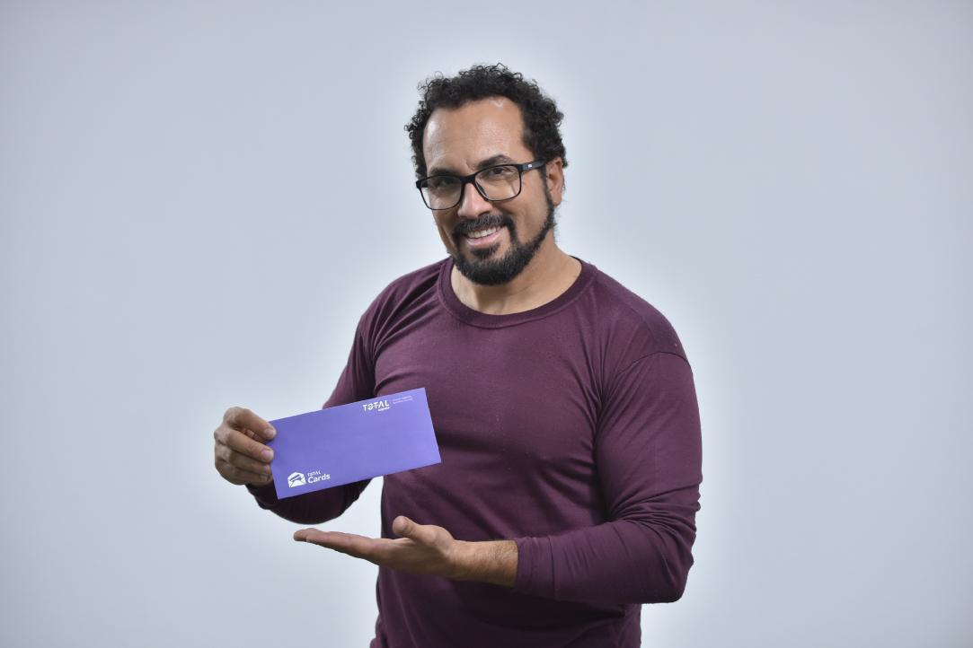 Um homem de cabelos cacheados, barba e óculos, vestindo uma camisa marrom de mangas compridas, segura e aponta para um envelope roxo com um sorriso. O fundo é totalmente branco.