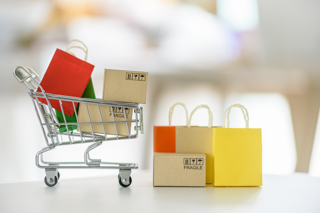 Um pequeno carrinho de compras cheio de caixas de papelão e sacolas coloridas fica sobre uma superfície branca. Ao lado, mais caixas de papelão com a etiqueta “FRÁGIL” são cercadas por sacolas de compras vermelhas, amarelas e laranja, representando um conceito de entrega de compras online.