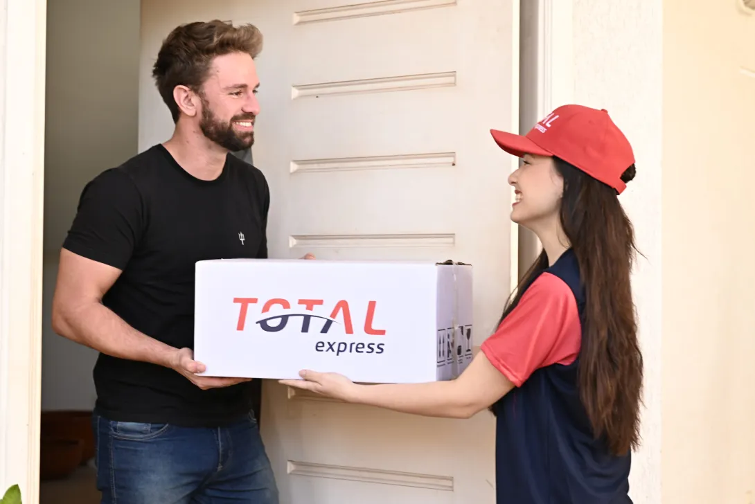 Um entregador de boné vermelho e uniforme azul entrega um grande pacote branco com o rótulo "TOTAL express" para um homem sorridente de camisa preta em sua porta.