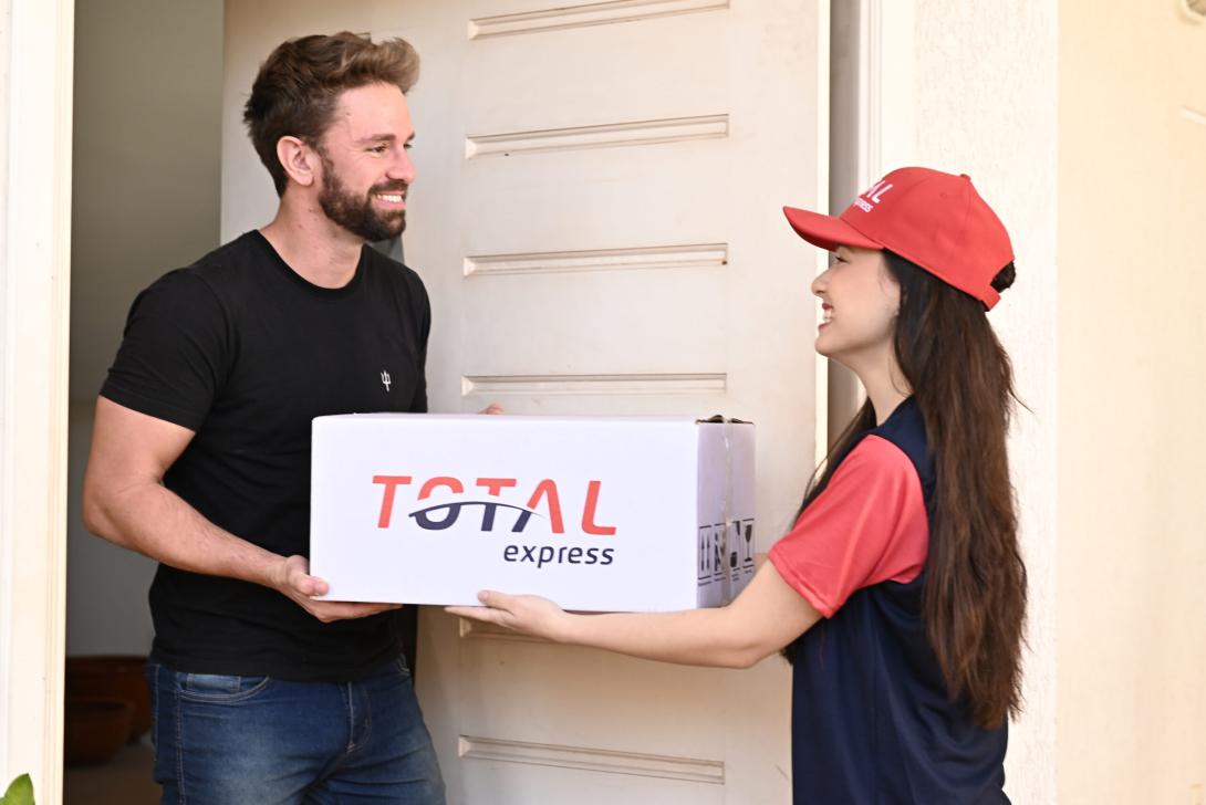 Um entregador de boné vermelho e uniforme azul entrega um grande pacote branco com o rótulo "TOTAL express" para um homem sorridente de camisa preta em sua porta.