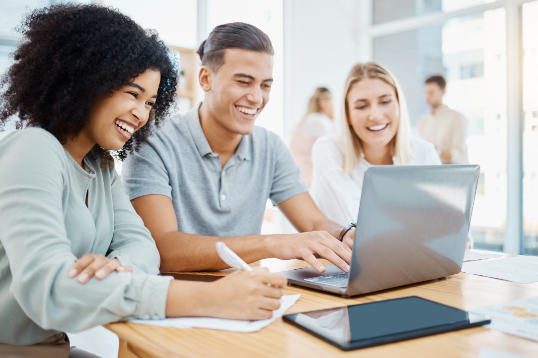 Três pessoas sorridentes sentadas ao redor de uma mesa com um laptop, colaborando e fazendo anotações. Eles parecem estar em um ambiente de escritório moderno e bem iluminado, com grandes janelas ao fundo. Papéis e um tablet também ficam visíveis sobre a mesa.