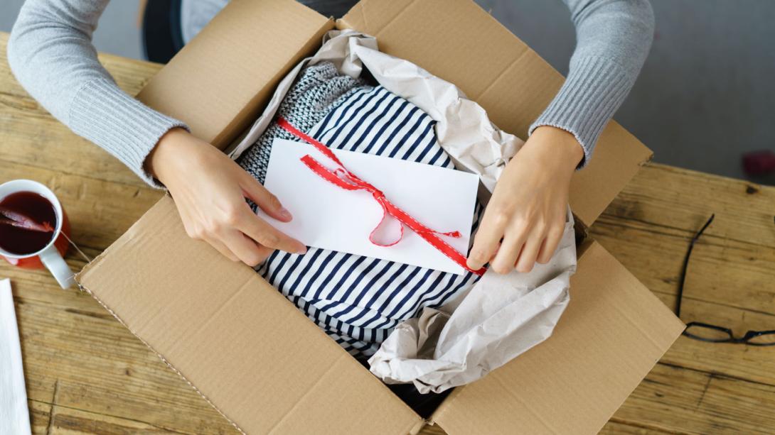 Uma pessoa abre uma caixa de papelão contendo um presente de tecido listrado, amarrado com uma fita vermelha e um envelope branco por cima. A caixa está cercada por papel de embalagem amassado sobre uma mesa de madeira. Uma xícara de chá está visível ao lado.
