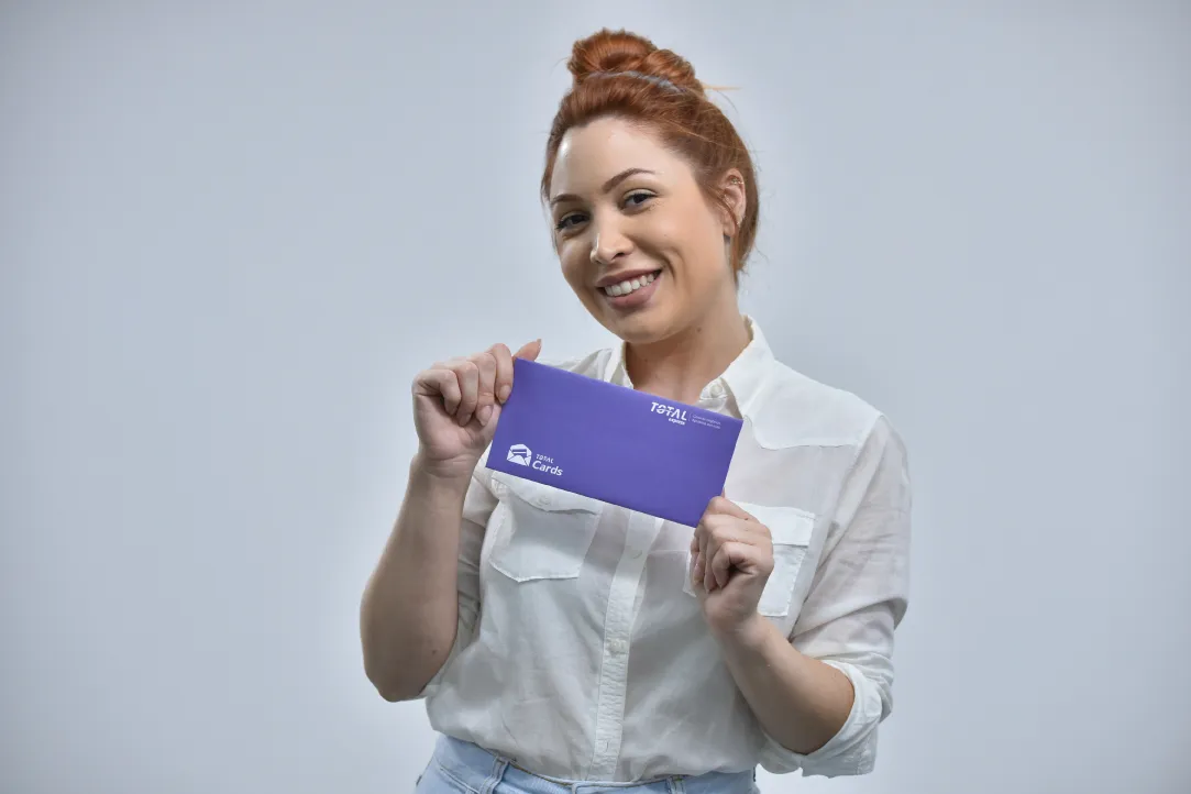 Uma pessoa com penteado em coque está sorrindo e segurando um envelope roxo com as duas mãos. Eles estão vestindo uma camisa branca de botões e contra um fundo cinza claro. O envelope contém texto e logotipos em branco.
