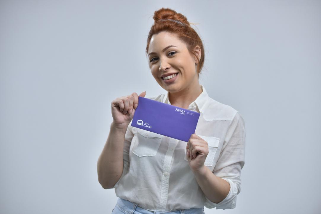 Uma pessoa com penteado em coque está sorrindo e segurando um envelope roxo com as duas mãos. Eles estão vestindo uma camisa branca de botões e contra um fundo cinza claro. O envelope contém texto e logotipos em branco.