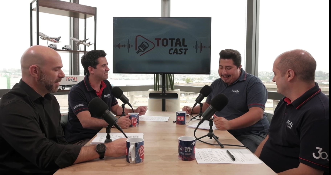 Quatro homens estão sentados ao redor de uma mesa com microfones, conversando, gravando um podcast intitulado “Total Cast”. O fundo apresenta uma grande janela com vista externa, uma tela com a logomarca do podcast e uma estante com itens decorativos.