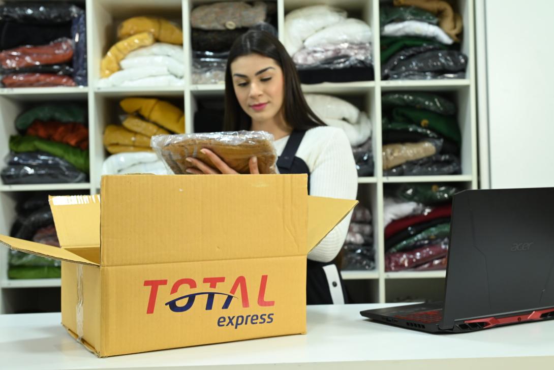 Uma mulher está atrás de um balcão, examinando uma peça de roupa embalada ao lado de uma caixa aberta do Total Express. Ao fundo, prateleiras com roupas dobradas. Um laptop está aberto no balcão.