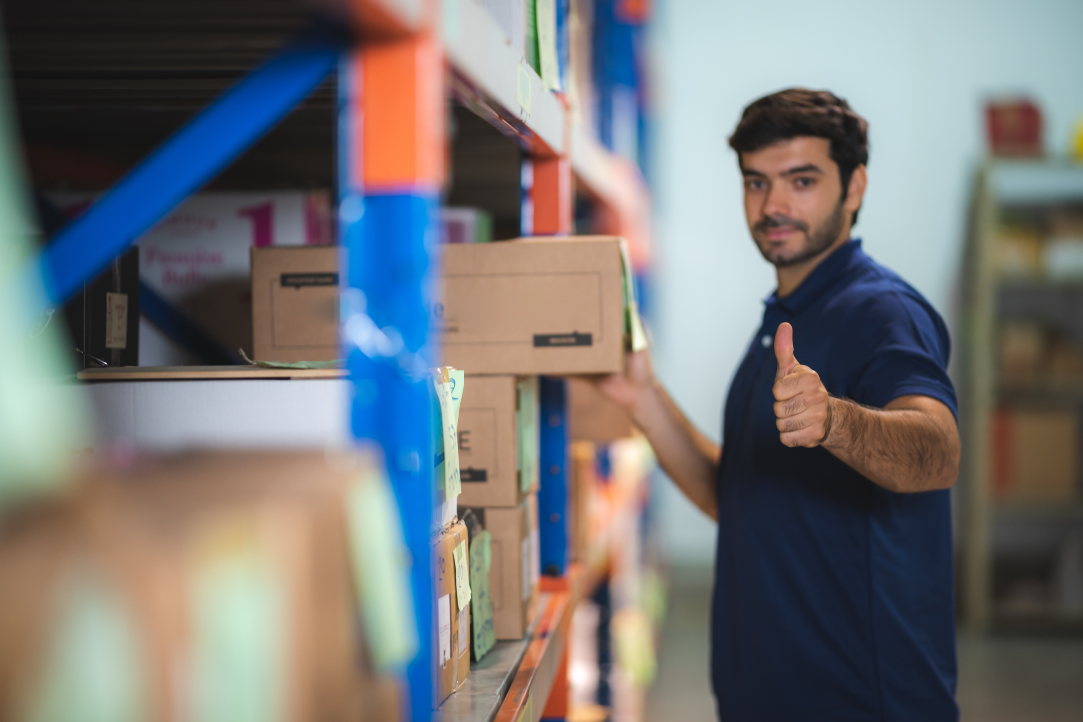 Um homem de camisa pólo azul está em um armazém, organizando caixas em uma prateleira. Ele faz um sinal de positivo com a mão direita enquanto olha para a câmera, indicando satisfação ou aprovação. Vários rótulos e embalagens coloridas são visíveis ao seu redor.
