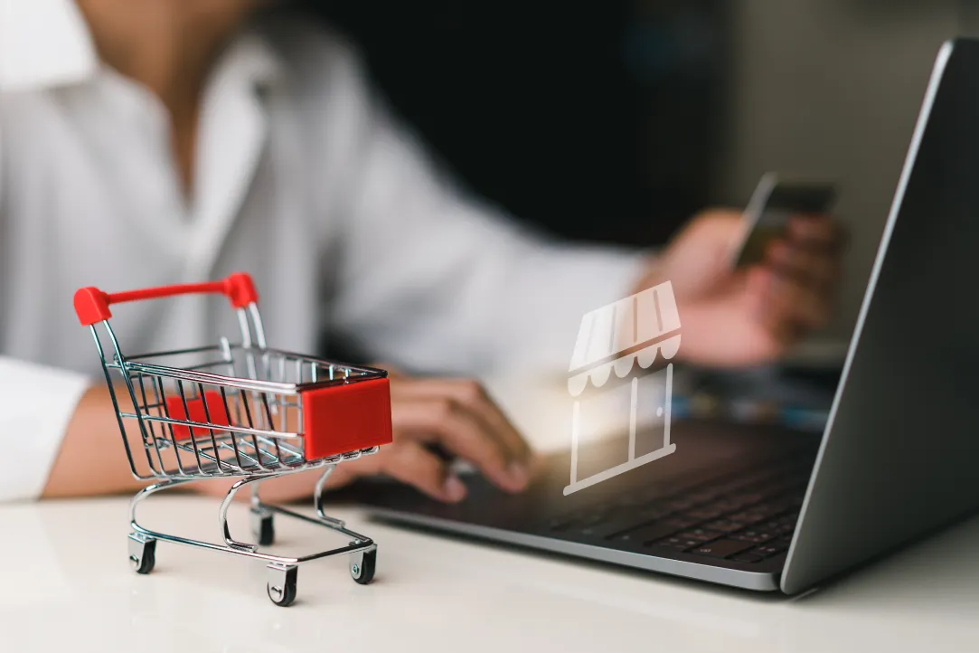 Uma pessoa segurando um cartão de crédito com uma das mãos e usando a outra para digitar no teclado de um laptop. Um pequeno carrinho de compras de brinquedos é colocado na mesa próxima. Um ícone de uma loja aparece na tela do laptop, representando compras online.