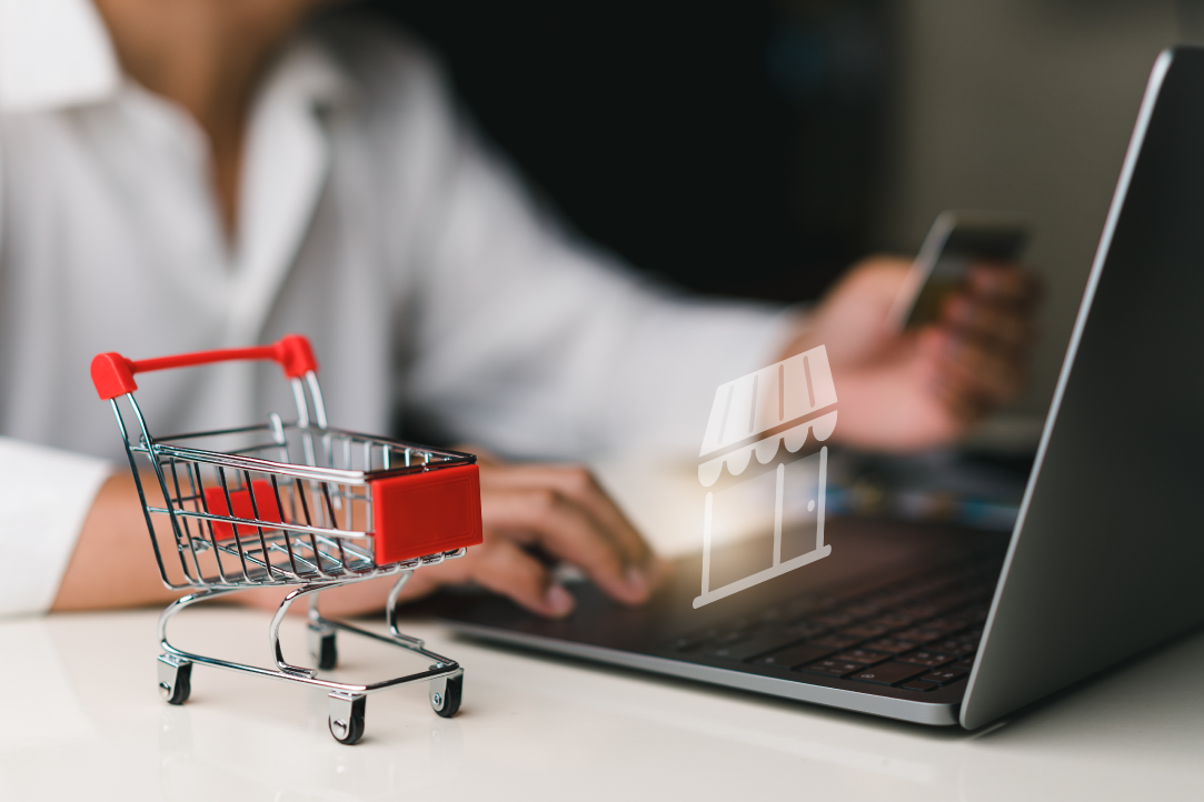 Uma pessoa segurando um cartão de crédito com uma das mãos e usando a outra para digitar no teclado de um laptop. Um pequeno carrinho de compras de brinquedos é colocado na mesa próxima. Um ícone de uma loja aparece na tela do laptop, representando compras online.