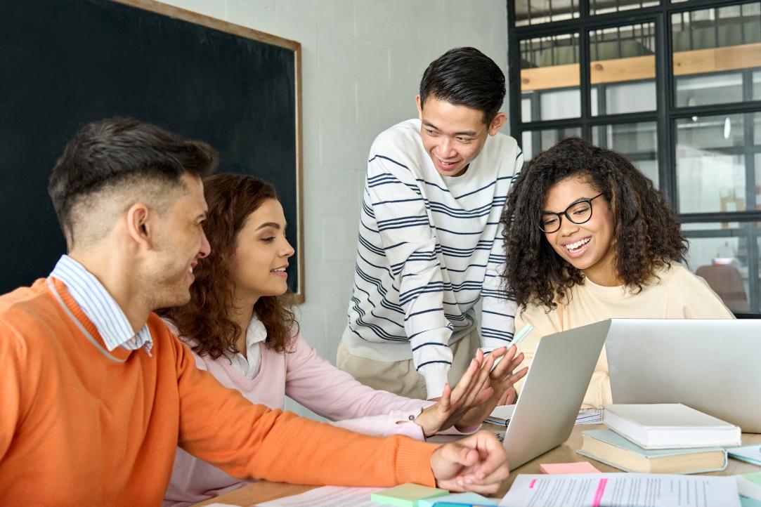 Um grupo de quatro colegas diversos está reunido em torno de uma mesa, envolvidos em uma discussão animada. Duas mulheres e dois homens estão sorrindo e interagindo entre si, com uma mulher usando um laptop. O cenário parece ser um escritório casual ou uma sala de reuniões.