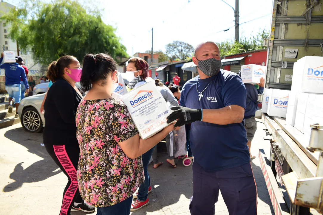 Várias pessoas usando máscaras distribuem e recebem caixas de mantimentos de um caminhão em uma rua. Um homem de camisa azul e luvas entrega uma caixa com a inscrição "Domilici" para uma mulher de camisa floral. Outros indivíduos ficam próximos com caixas.