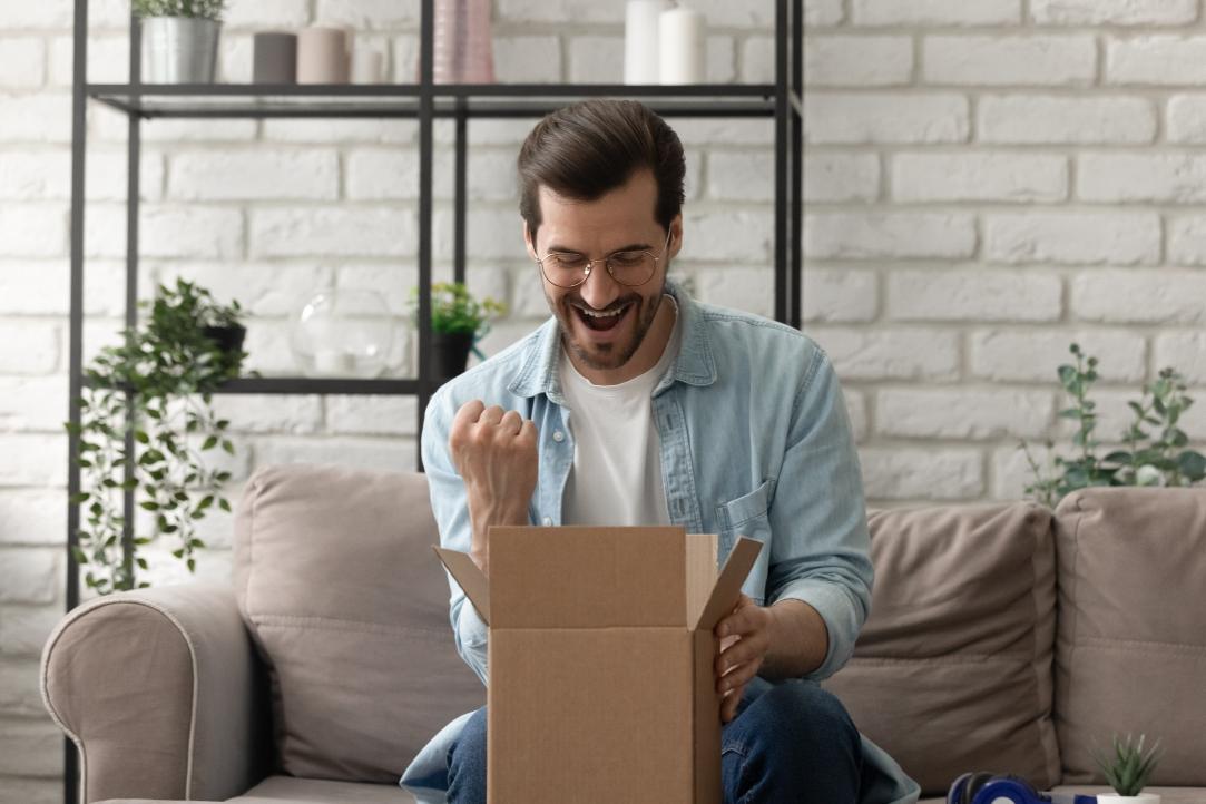 Um homem de óculos e barba, vestindo uma camisa azul clara sobre uma camiseta branca, está sentado em um sofá em frente a uma caixa de papelão aberta, parecendo animado e cerrando o punho em triunfo. Prateleiras com plantas e decoração ficam ao fundo.
