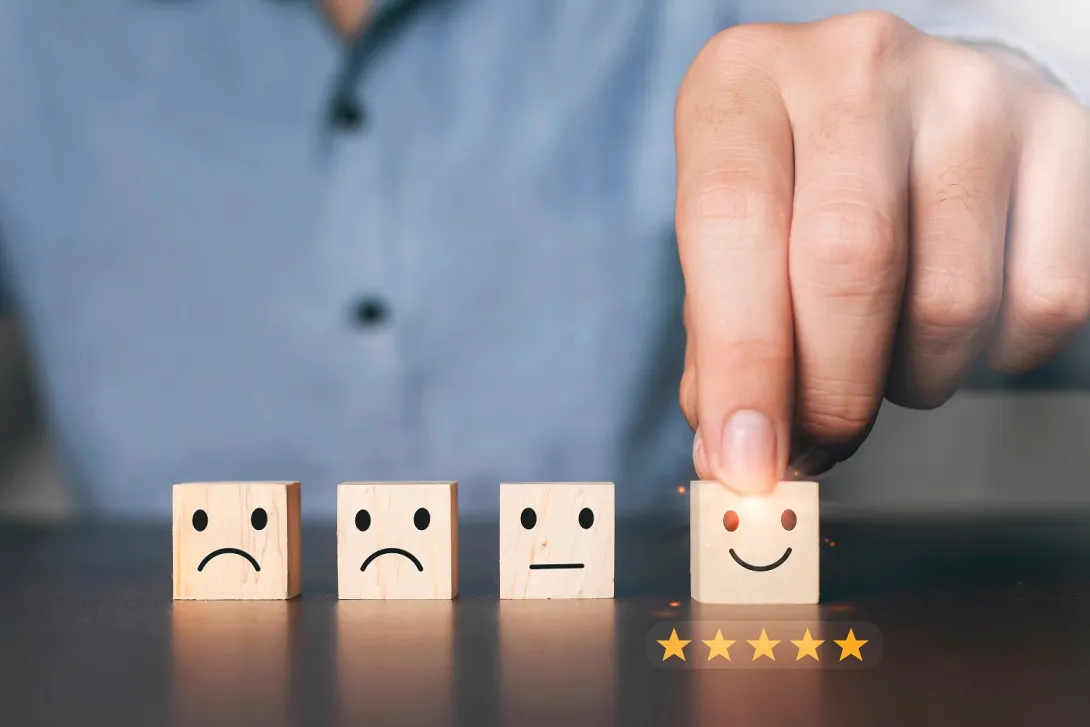 Uma pessoa organiza quatro blocos de madeira, cada um exibindo emoticons diferentes: triste, neutro, levemente sorridente e muito feliz. A pessoa toca o bloco muito feliz, com cinco estrelas douradas abaixo, significando excelente avaliação ou satisfação.