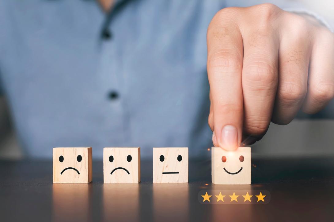 Uma pessoa organiza quatro blocos de madeira, cada um exibindo emoticons diferentes: triste, neutro, levemente sorridente e muito feliz. A pessoa toca o bloco muito feliz, com cinco estrelas douradas abaixo, significando excelente avaliação ou satisfação.