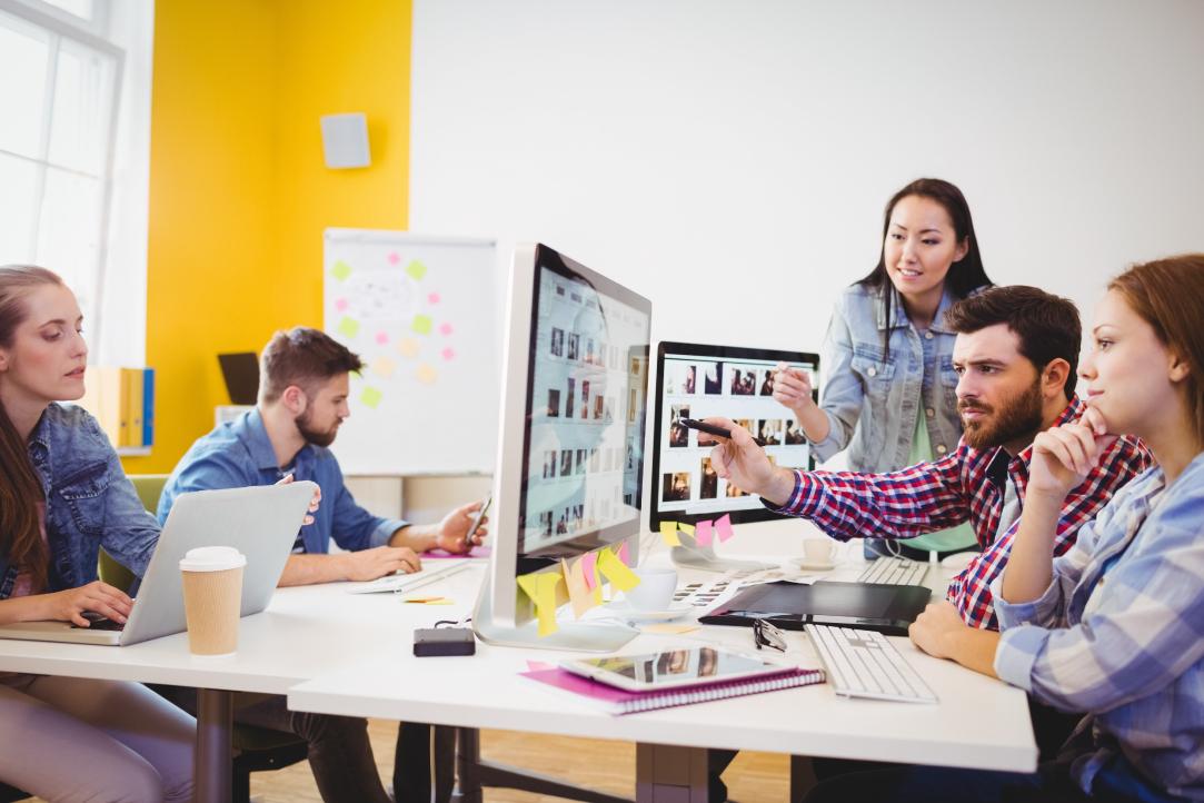 Um grupo de cinco pessoas trabalhando em um escritório moderno. Três indivíduos estão reunidos em torno de duas telas de computador, discutindo algo atentamente. Outros dois estão trabalhando em laptops em uma mesa próxima. A sala tem paredes amarelas e brancas brilhantes e uma janela com luz natural.