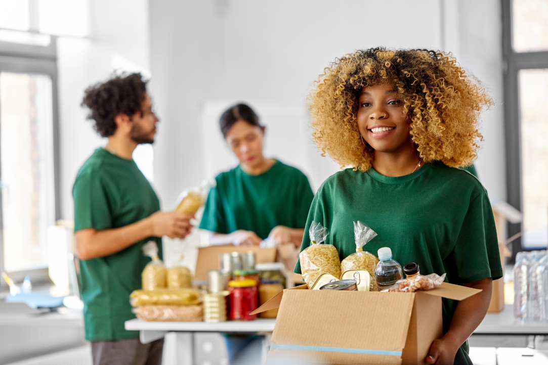 Uma mulher sorridente com cabelos cacheados segura uma caixa cheia de vários alimentos, incluindo massas e enlatados. Ao fundo, outras duas pessoas vestindo camisas verdes separam itens semelhantes sobre uma mesa. Eles parecem estar trabalhando como voluntários em um banco de alimentos.