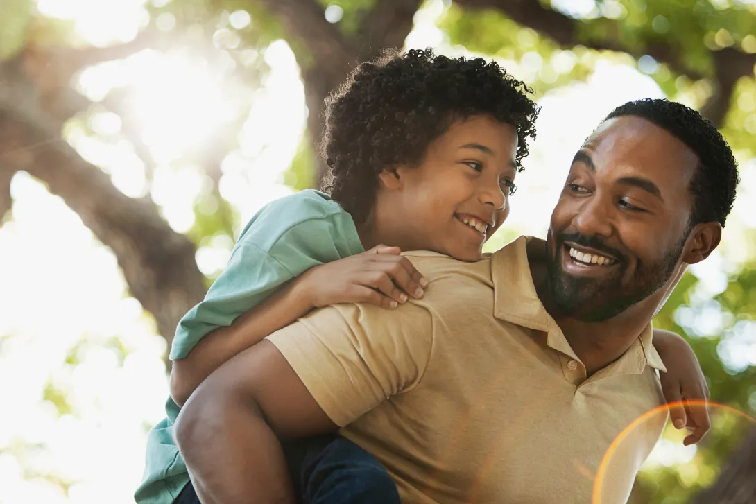 Filho e pai pretos abraçados e sorrindo, a criança está com verde e o pai com camiseta marrom, o fundo contém uma árvore. 