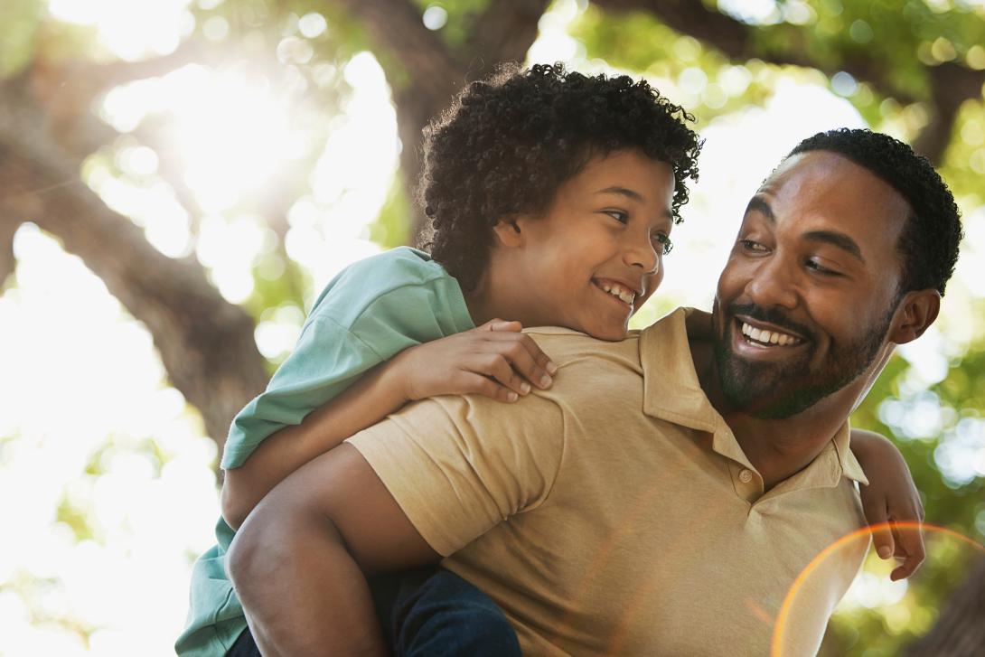 Filho e pai pretos abraçados e sorrindo, a criança está com verde e o pai com camiseta marrom, o fundo contém uma árvore. 