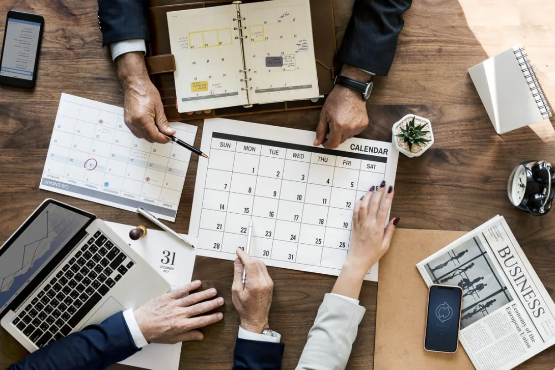 Na imagem vemos várias mãos em cima de uma mesa mexendo no calendário comercial.