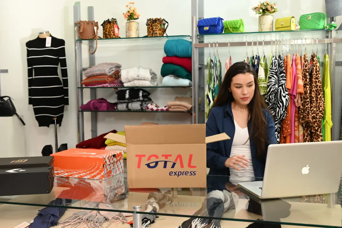 Empresária gerenciando pedidos e logística no computador, com caixas da Total Express, representando a gestão de e-commerce.