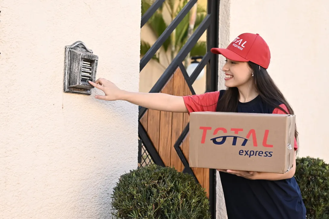 Uma pessoa tocando a campainha de uma casa para realizar uma entrega. Nas mãos, vemos uma caixa com a logo da Total Express.