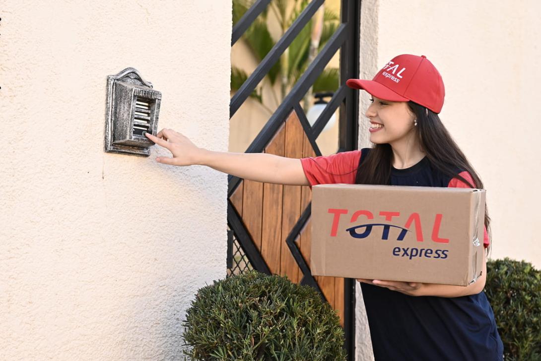 Uma pessoa tocando a campainha de uma casa para realizar uma entrega. Nas mãos, vemos uma caixa com a logo da Total Express.