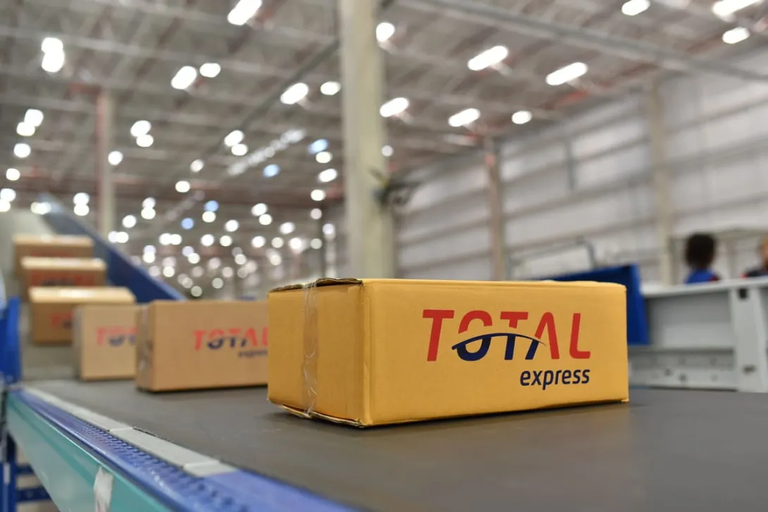 Caixa com o logo da Total Express em uma esteira de distribuição, ilustrando o processo de supply chain em um centro de logística.