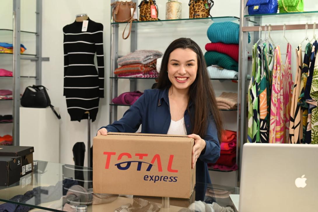Jovem empreendedora sorridente segura uma caixa de entrega em sua loja de moda, ilustrando a etapa de envio e a praticidade de como vender roupas na internet.