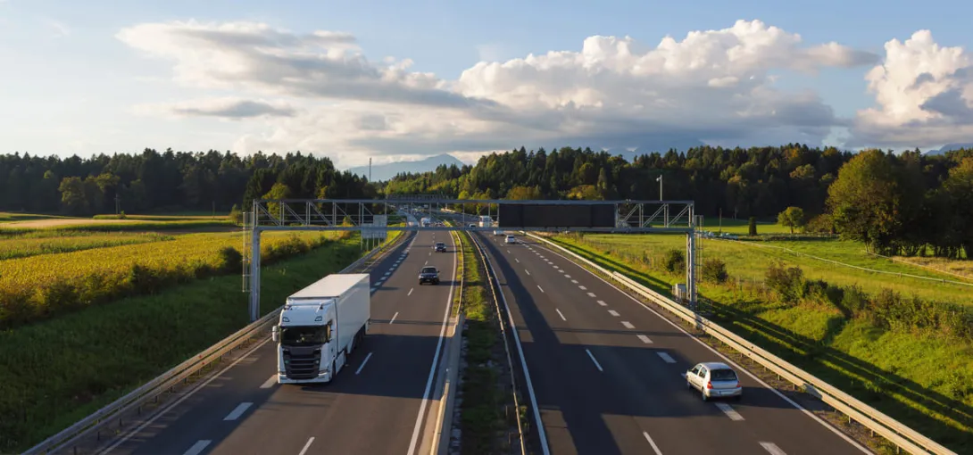 Um caminhão branco de carga viaja em uma rodovia de várias faixas no final da tarde, passando por campos e florestas com montanhas distantes, exemplificando a infraestrutura e a logística do transporte doméstico.