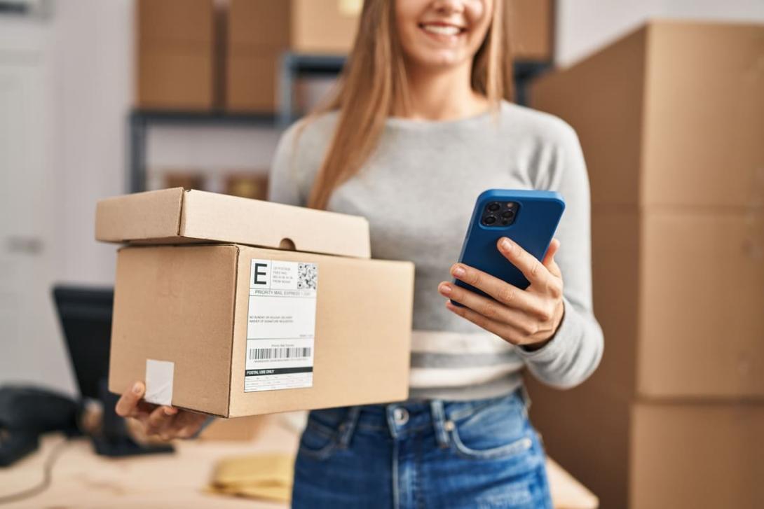 Mulher sorridente segura caixas de encomendas e um smartphone, representando a facilidade de exercer o direito de arrependimento em compras online.
