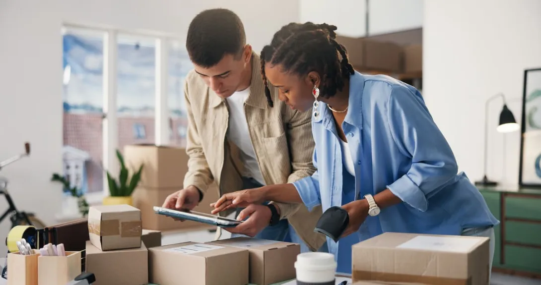 A gestão de pedidos no e-commerce é ilustrada por um casal de jovens empreendedores conferindo caixas de encomenda com um tablet e um leitor de código de barras em um escritório.