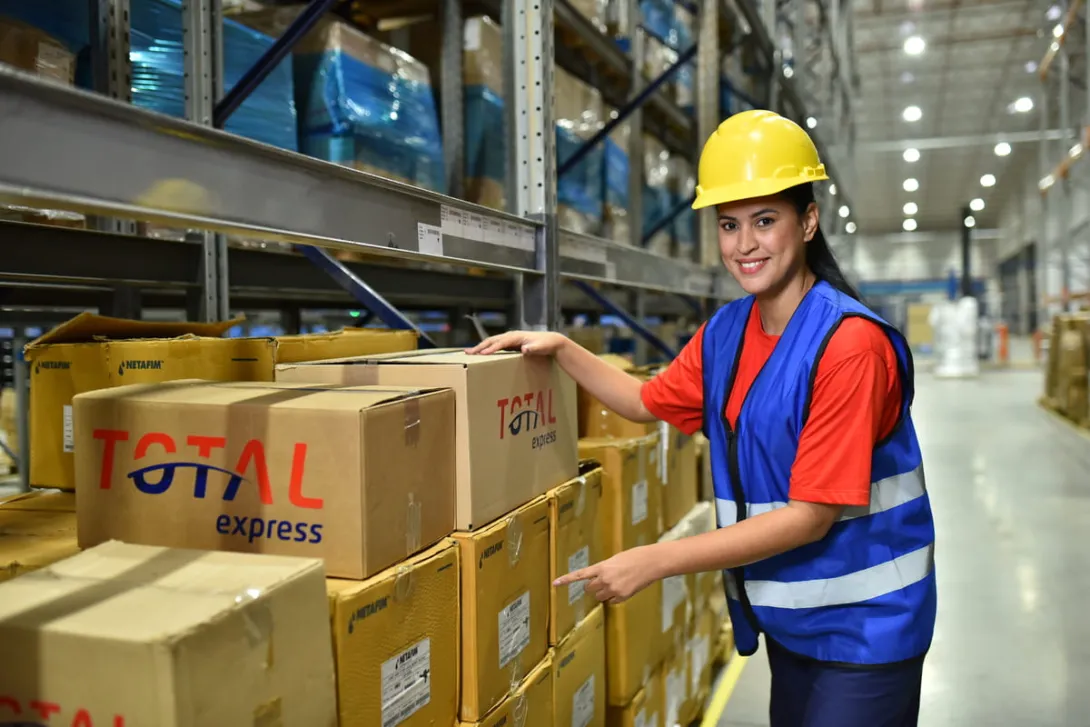Funcionária com capacete e colete de proteção organiza caixas da Total Express em um galpão de estoque, ilustrando a operação de fulfillment ou logística própria.