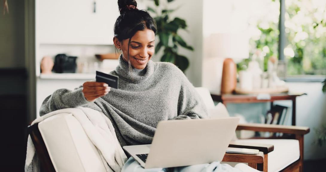 Mulher sorridente sentada no sofá usa um notebook e segura um cartão de crédito, ilustrando as novas tendências em meios de pagamento.