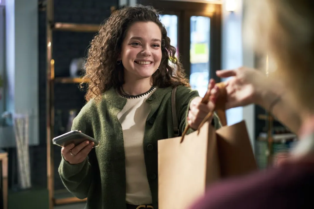 Mulher sorridente com celular recebe sacola de compras em loja no Dia do Cliente.